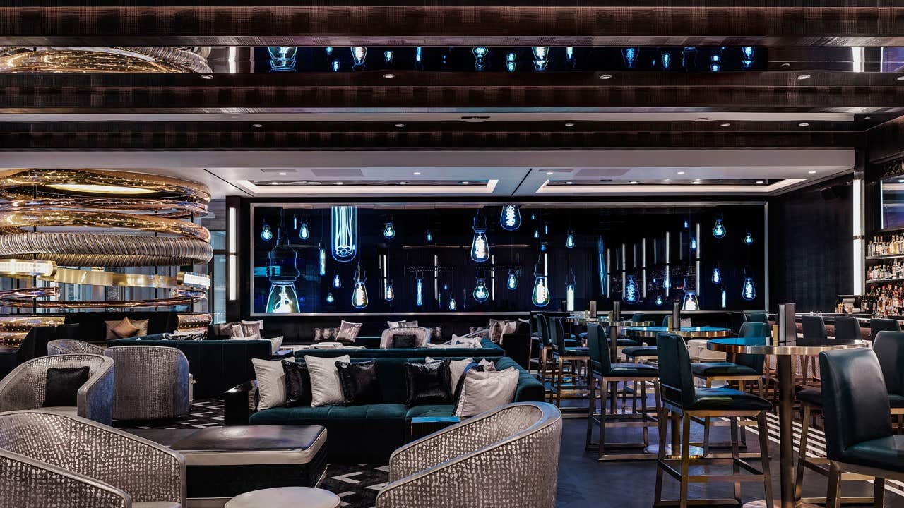 Interior of a sleek, modern bar with gold bracelet wall feature, silver armchairs and green velvet couches.