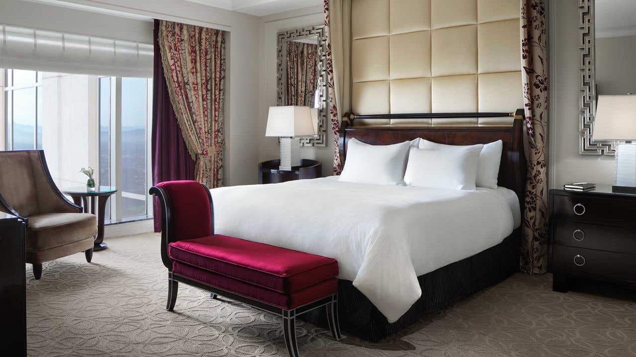 Hotel room with a king-sized bed with white linens and a plush headboard, flanked by nightstands with lamps and a red bench at the foot of the bed. A window is on the left with an armchair and side table in front of it.  