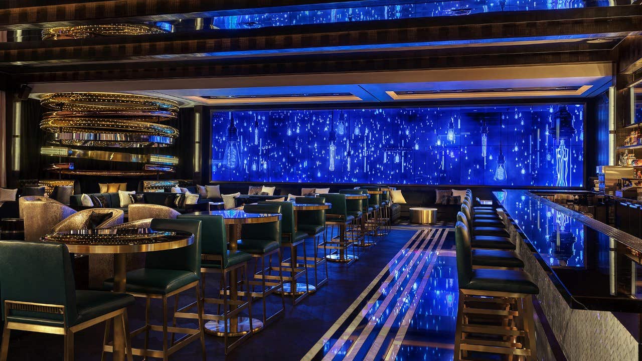 Interior of a sleek, modern bar with dark marble, gold accents and a large video wall showcasing a blue background covered in Edison bulbs.