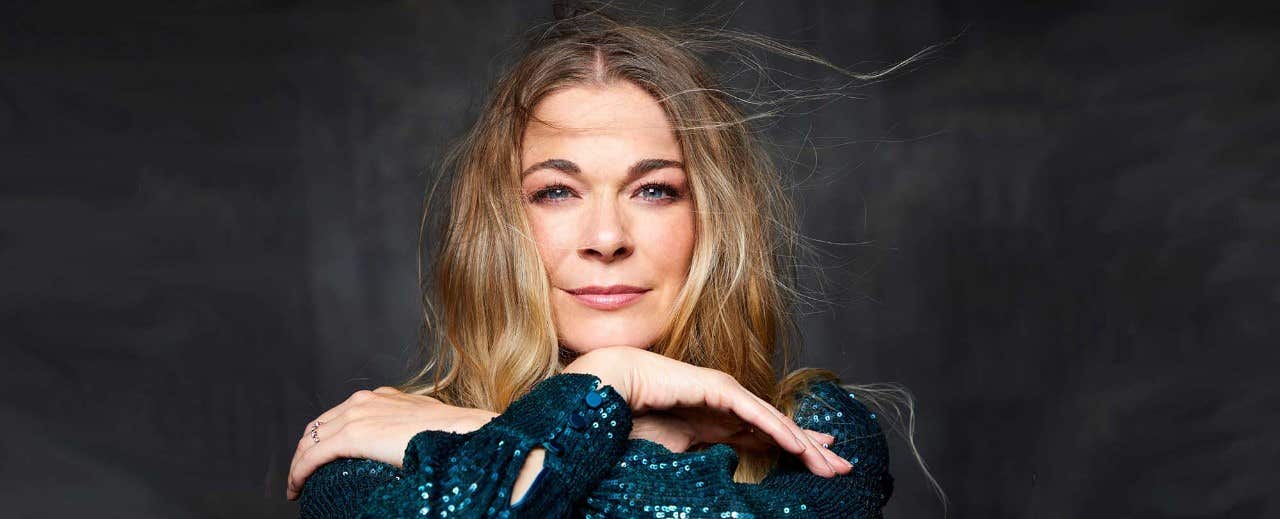 Woman with long blond hair in a sequined green outfit posing with her arms crossed under her chin against a dark background.