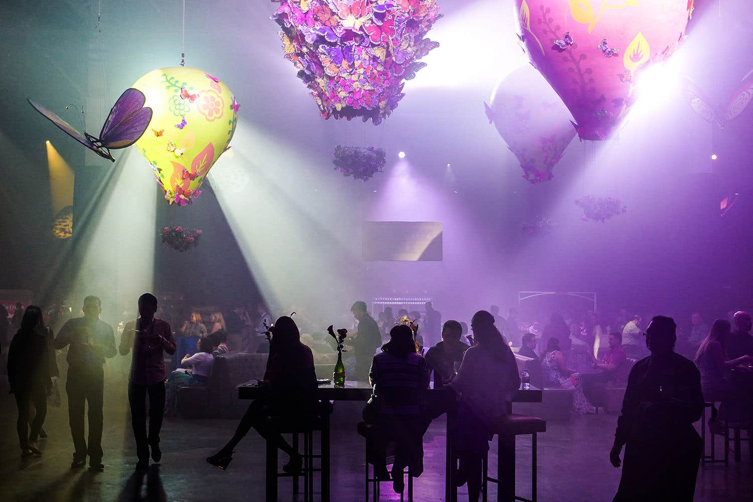 A dimly lit event at The Venetian in Las Vegas with people socializing, featuring large, illuminated balloon decorations with floral designs and beams of light piercing through a hazy room.