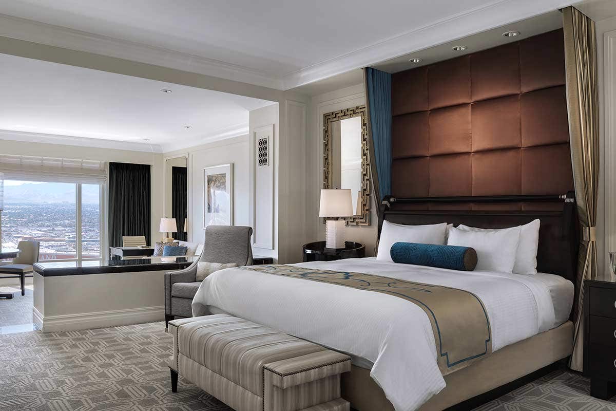 Luxurious hotel room with a king-sized bed, seating area, desk, and large windows offering a city view. Modern decor.