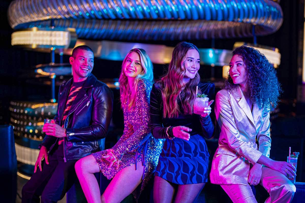 Four friends in fashionable outfits smiling and holding drinks at a vibrant nightlife venue.